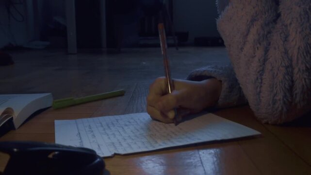 In a dimly lit room, a hand captures thoughts on paper with a pen. The scene features a cozy blue robe and wooden flooring, embodying the quiet essence of late-night reflection and creative focus.