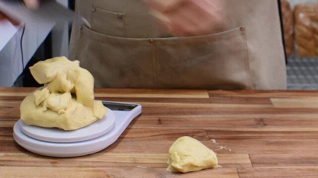 Close-up of a man&rsquo;s hands weighing, cutting, and shaping dough into balls on a scale on a wooden tabletop kitchen, showing artisanal bread preparation and baking technique