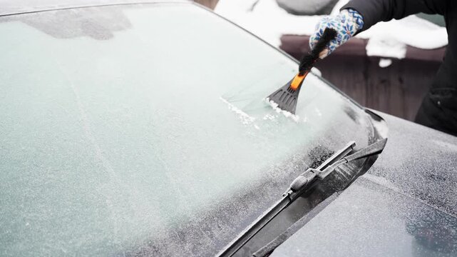 Wide view of a frosted vehicle windshield as a gloved hand clears a frozen patch near the wipers with an orange brush tool, improving visibility before a cold morning drive.