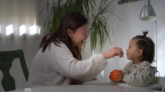 Lifestyle clip of an Asian mother and her half Asian European child sitting at the table at home, playfully sticking out their tongues while the child holds an orange, sharing a joyful moment.