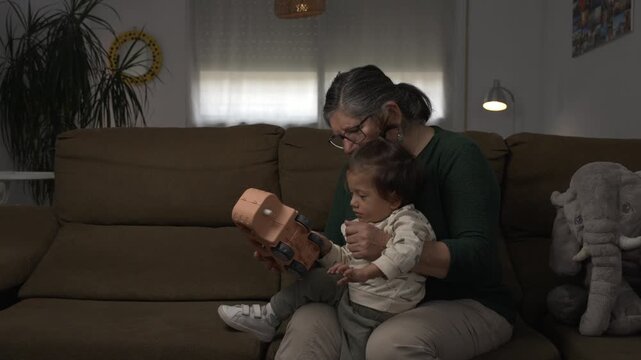 Indoor lifestyle scene of a Spanish senior woman and a young Asian child sitting on a sofa and playing together with a toy truck, spinning its wheels in a calm and affectionate moment at home