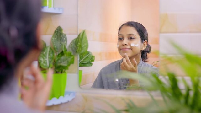 Young female applying sunscreen on her face in front of a mirror during morning skincare routine