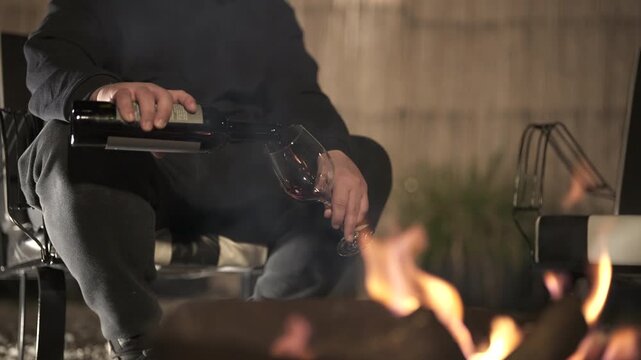 Person relaxes near firewood and flames while pouring wine, calm evening setting