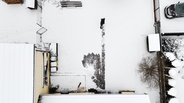 Top-down drone view of a person shoveling a narrow path through fresh snow, revealing cobblestone in a residential backyard during winter after a storm.