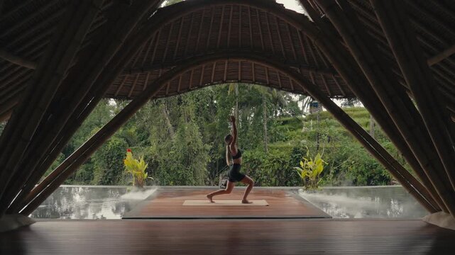 Blonde caucasian woman doing warrior yoga pose in bamboo yoga shala with jungle view