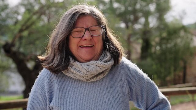 A senior woman smiles and gradually starts laughing shyly as the camera slowly moves toward her in a calm outdoor setting.