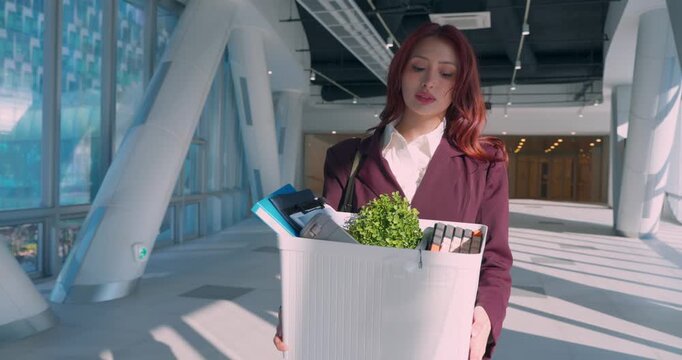 A worried young Latin professional in a maroon suit walks through a bright modern office lobby carrying a cardboard box with her plant and belongings following a corporate layoff