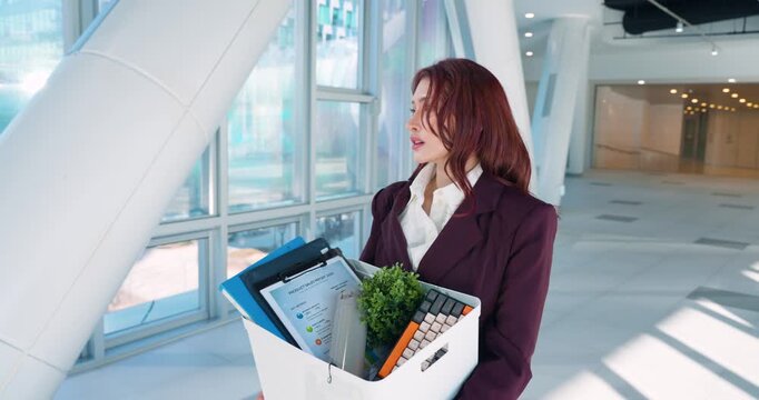 Sad worried young woman in maroon suit carries cardboard box with belongings plant and documents walking through bright glass office atrium after layoff