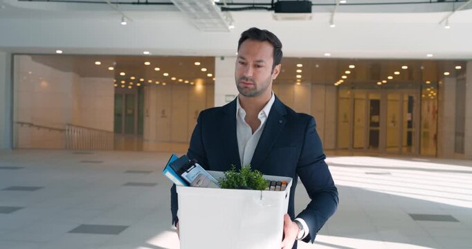 Stressed man in suit holds cardboard box with belongings plant and documents walking through bright glass office atrium after corporate layoff slow motion
