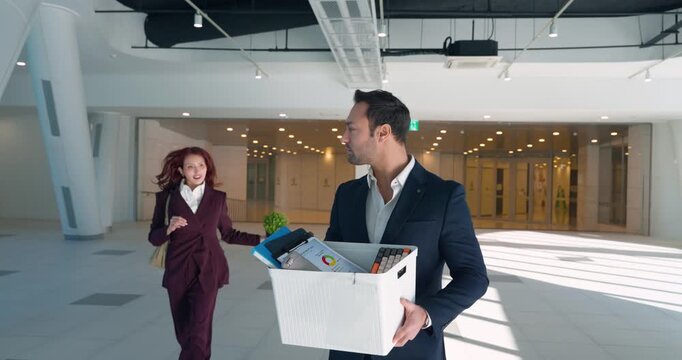 Sad businessman in suit holds cardboard box of belongings walking modern office while cold female colleague throws plant into box and walks away indifferent