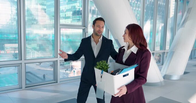 Businessman in suit comforts worried young woman holding box of belongings after being fired while walking together in modern glass office atrium