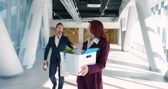 A worried young woman walks through a modern glass office lobby carrying a box of personal items after being laid off; man coworker follows to return her keyboard and say final goodbye - tracking shot