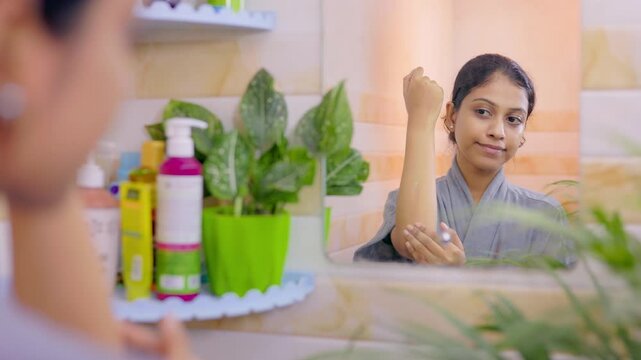 Young lady applying hydrating body lotion on her forearm to prevent dryness, standing in front of a bathroom mirror.