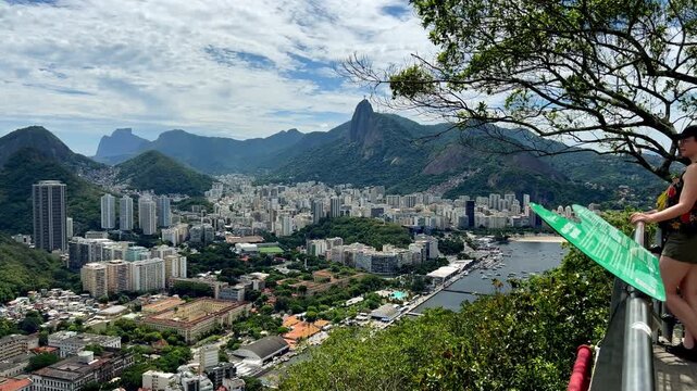 A traveler stands at a railing on Urca Hill, looking out over the sprawling city of Rio de Janeiro toward Corcovado Mountain and the Christ the Redeemer statue under a dramatic sky.