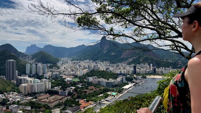 A traveler stands at a railing on Urca Hill, looking out over the sprawling city of Rio de Janeiro toward Corcovado Mountain and Christ the Redeemer under a dramatic sky.