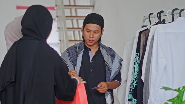 Young Muslim man serving customers and using EDC machine for payment transaction in boutique