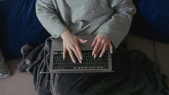 Close-up view of a young woman typing on her laptop while sitting comfortably on a sofa at home. The cozy setting and soft blanket emphasize remote work and modern digital lifestyle.
