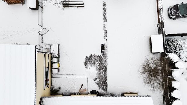 Top-down drone shot of a person clearing a cobblestone walkway in a snowy private courtyard, revealing dark stone patterns with wide white copy space.