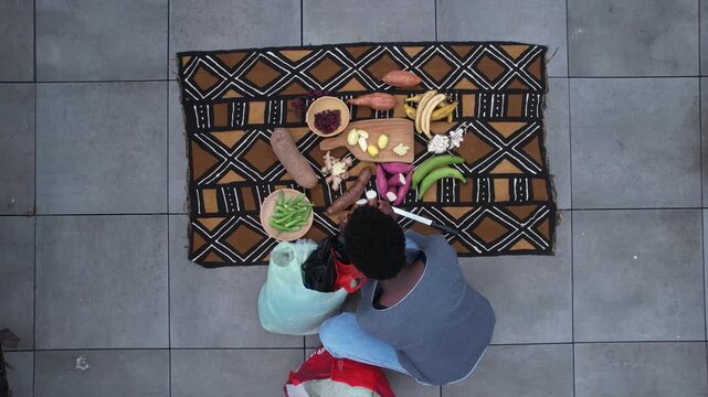 Woman preparing a flat lay of fresh produce on a patterned African textile on a tiled patio, with multiple terracotta pots of herbs and seedlings arranged to the side in daylight.
