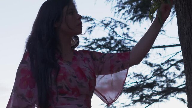 A young woman in a floral dress gently touches tree leaves in Melbourne's Croydon Park. The tranquil scene is illuminated by soft lighting, capturing an intimate, peaceful moment.