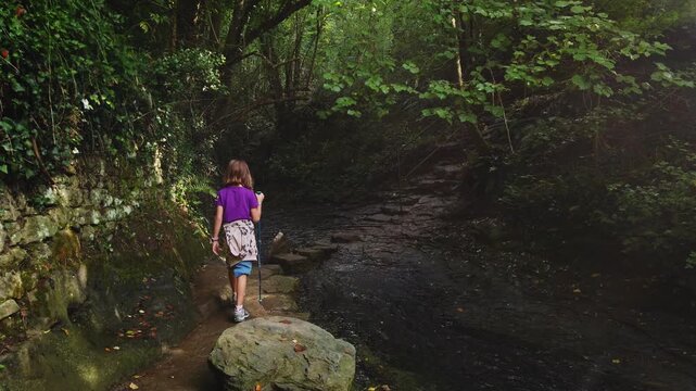 Young female explorer hiking with a pole along a scenic rocky path in a lush green forest in Rupit, Catalunya. This footage shows the concept of adventure, discovery, and active childhood