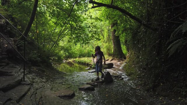 Adventurous little girl with a hiking stick carefully crossing a small river by stepping on stones in a lush green forest, exploring the natural beauty of Rupit, Barcelona, in Catalunya, Spain