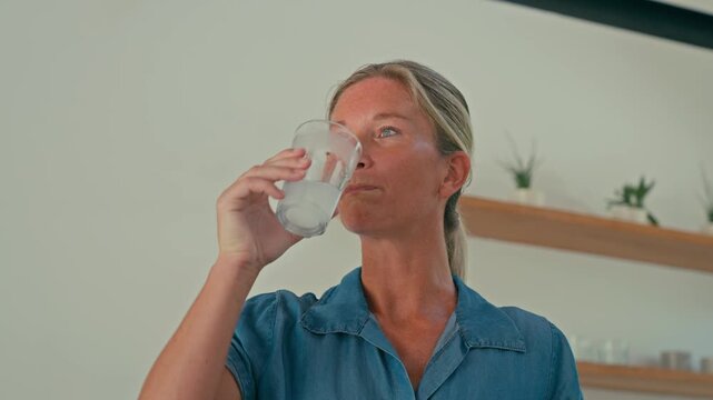 Fit blonde woman drinks creatine mixed with water from a glass in her kitchen at home, showing a healthy daily fitness routine