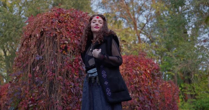 Red haired boho witch beckoning toward camera in vibrant autumn setting with colorful fall foliage, subtle Halloween mood, cinematic lateral tracking shot to the right, vertical format.