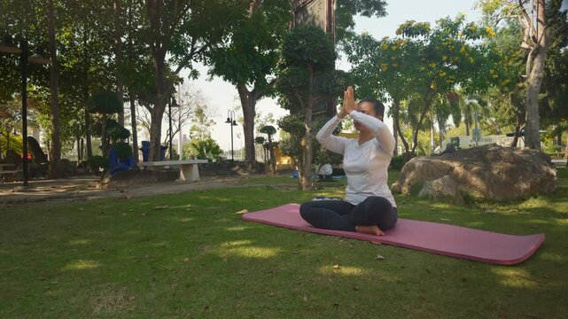 Young woman practicing morning meditation outdoors, raising hands with deep inhale before sitting calmly with eyes closed. Concept of mindfulness, breathing exercise and mental wellbeing.