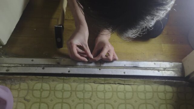 A top down POV shot of a woman carefully setting a nail into a doorway threshold. Precise initial hammering movement during a domestic repair project on a wooden parquet floor in a flat.