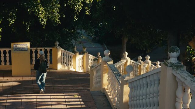 Woman walking past Plaza Nelson Mandela sign by a sunlit staircase in a lush Andalusian park, Almu&ntilde;&eacute;car, Spain.