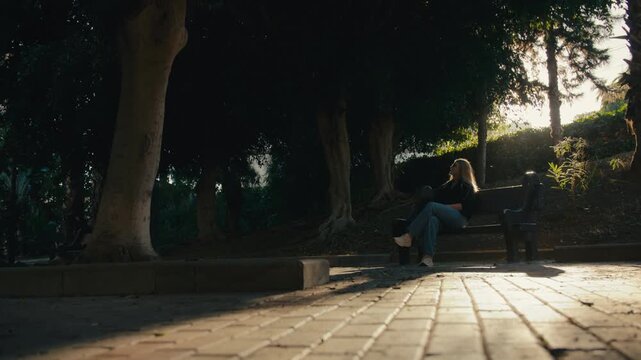 Young Scandinavian woman relaxing on a bench in a sunlit park surrounded by trees in southern Spain.