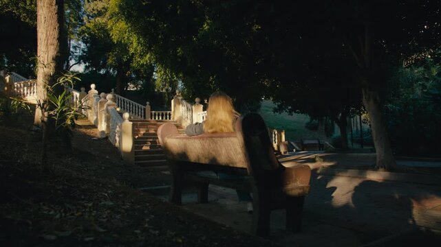 Young woman sitting on a bench in a shaded Andalusian park, watching a sunlit staircase at golden hour.