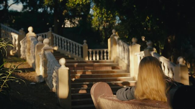Young woman resting on a bench facing sunlit staircase in a tranquil Andalusian park at golden hour.
