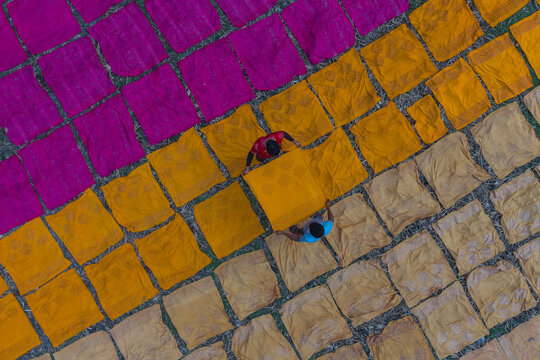 Narayanganj, Bangladesh - 08 March 2026: Aerial view of vibrant fabrics drying under the sun, creating a colorful patchwork with workers amidst the geometric patterns.