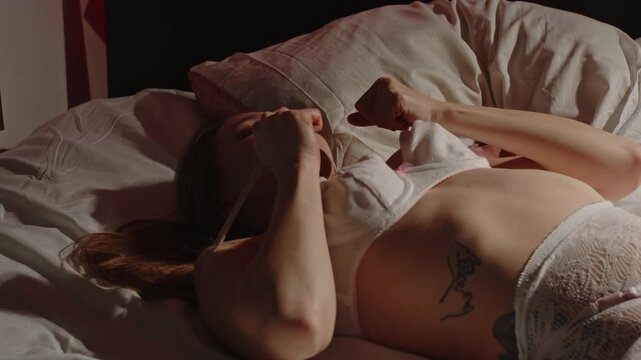 An attractive woman wearing enticing lingerie lies in bed and sensually plays with her brassiere in slow motion