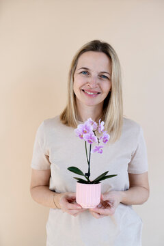A blonde woman wearing a brown shirt is holding an orchid and standing in front of a brown background