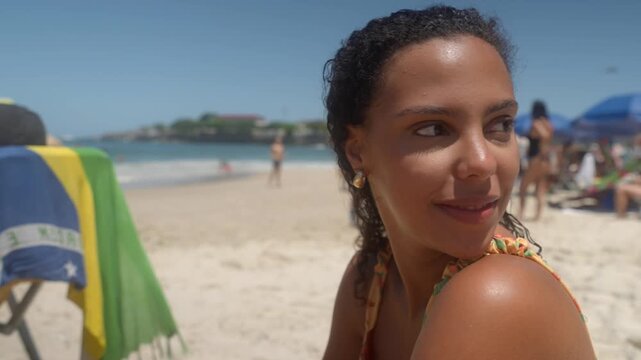 Young Latina woman with curly hair sipping a yerba mate tea from a plastic cup at Copacabana beach. Sun hits her face and shoulders. Static full shot during early afternoon, with blurred background.