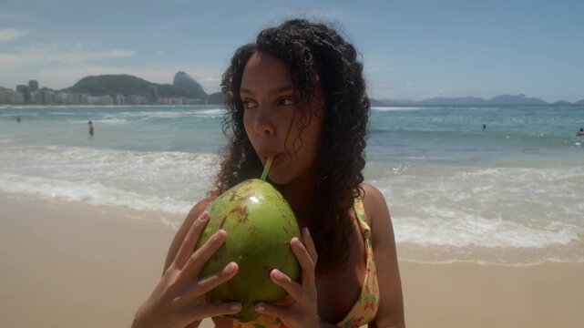 Young tanned woman standing at Copacabana beach holding and sipping from a green coconut and smiling at the camera. Static full shot with breaking waves and Sugar Loaf Mountain in background.