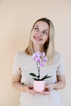 A blonde woman wearing a brown shirt is holding an orchid and standing in front of a brown background