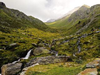 Obraz premium Alpine Valley with Stream and Waterfall Leading to a Mountain Pass Under Dramatic Clouds, Italian Alps Landscape