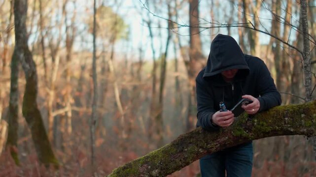 Slow motion. Young man in black hoodie vaping e-cigarette and texting on smartphone in forest. Male using mobile phone and smoking electronic cigarette outdoors among trees.