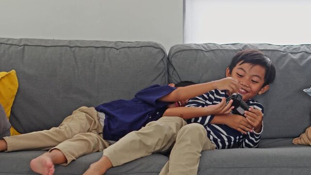 Two playful Asian brothers laughing and wrestling for a game controller on a gray sofa