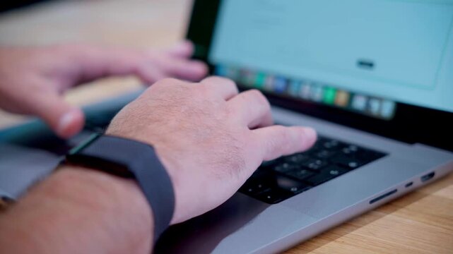 Male hands typing on a laptop keyboard focusing on a black smartwatch on the wrist. Professional working environment with shallow depth of field blurring the screen.