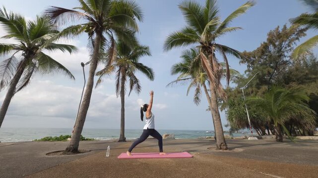Wide shot of woman stepping forward into Warrior pose, raising arms overhead and transitioning into Reverse Warrior during yoga flow on tropical beach by the ocean.