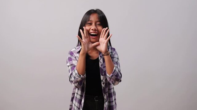 Friendly young Asian girl shouting or calling out loudly with cupped hands on a grey studio background