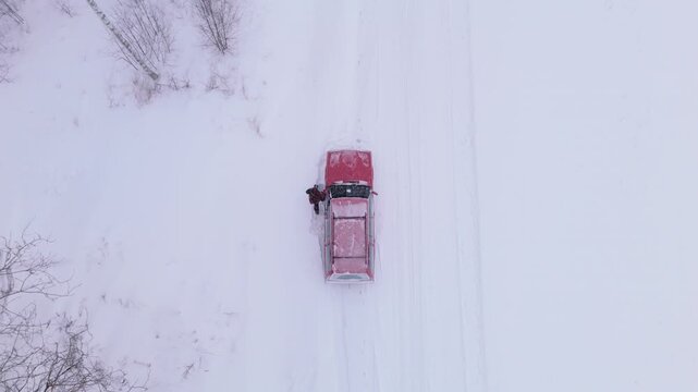 Man exits broken down red car next to snowy winter road in countryside, overhead