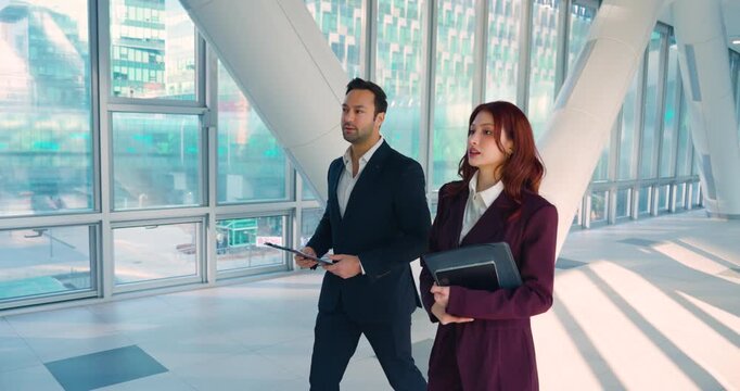 Confident business man and woman in suits walk discuss holding clipboard folder through spacious modern corporate lobby with glass walls slow motion