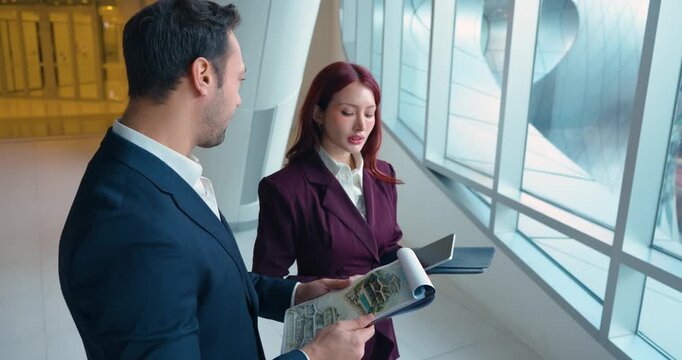 Smiling businessman and businesswoman architects happily discuss colorful architectural concept drawings and site plans in modern corporate lobby