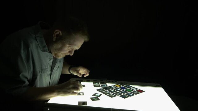 Male photo editor looks at colour slide film on lightbox in dark room
